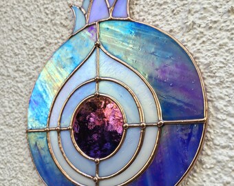 May include: A stained glass pomegranate in shades of blue, purple, and white, with iridescent effects. The design includes a central circular element with concentric rings and copper framing. A brown cord is attached for hanging. The piece is approximately 15 cm in diameter.