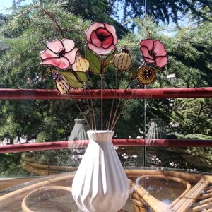 K&ouml;nnte beinhalten: Eine wei&szlig;e Keramikvase enth&auml;lt einen Strau&szlig; handgefertigter Glasblumen. Die Blumen haben rosa und rote Bl&uuml;tenbl&auml;tter, gr&uuml;ne Bl&auml;tter und Details von Samenkapseln. Die Anordnung befindet sich auf einem Tisch.