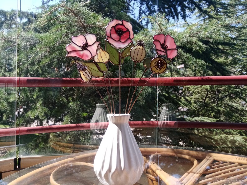 K&ouml;nnte beinhalten: Eine wei&szlig;e Keramikvase enth&auml;lt einen Strau&szlig; handgefertigter Glasblumen. Die Blumen haben rosa und rote Bl&uuml;tenbl&auml;tter, gr&uuml;ne Bl&auml;tter und Details von Samenkapseln. Die Anordnung befindet sich auf einem Tisch.