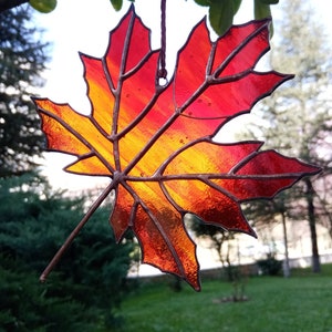 May include: A stained glass maple leaf with a gradient of red, orange, and yellow. The leaf is hanging from a string and is in front of a green grassy background.