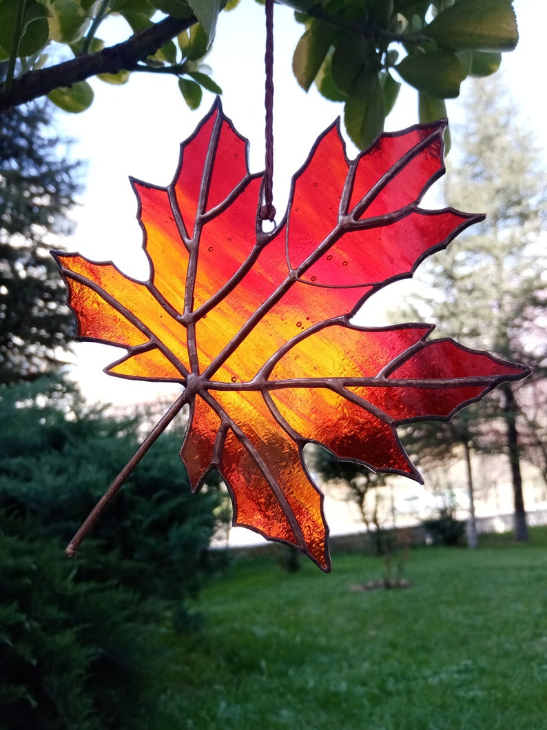 May include: A stained glass maple leaf with a gradient of red, orange, and yellow. The leaf is hanging from a string and is in front of a green grassy background.