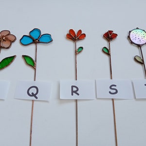 May include: Five colorful, handcrafted glass flower garden stakes. Each stake features a different flower design in shades of peach, turquoise, orange, red, and iridescent. The flowers are mounted on copper stems with green glass leaves. White cards below each flower are labeled with the letters P, Q, R, S, and T.