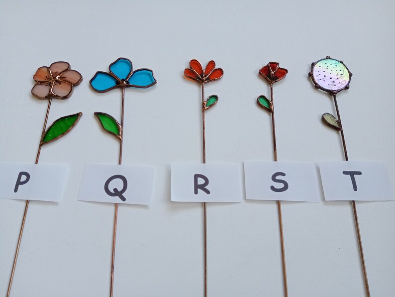 May include: Five colorful, handcrafted glass flower garden stakes. Each stake features a different flower design in shades of peach, turquoise, orange, red, and iridescent. The flowers are mounted on copper stems with green glass leaves. White cards below each flower are labeled with the letters P, Q, R, S, and T.