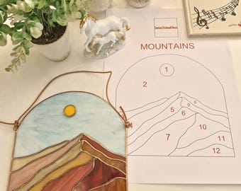 May include: A stained glass mountain scene with a blue sky and yellow sun. The mountain is made of various earth-toned glass pieces. The piece is framed with a copper-coloured metal. A paper with the word "MOUNTAINS" and a numbered mountain outline is next to the glass.