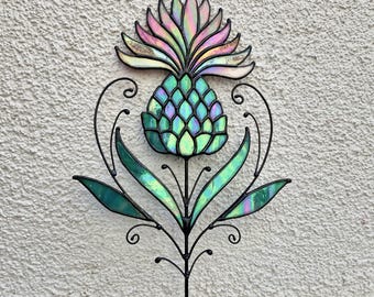 May include: A decorative metal and stained glass thistle garden stake. The thistle features a teal and iridescent pineapple-like head, green leaves, and a black metal stem with decorative swirls. The top of the thistle has pink and iridescent petals.