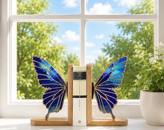 May include: A pair of bookends featuring stained glass butterfly wings in shades of blue with gold-toned metal edges, mounted on rectangular wooden bases, supporting a single book with a white spine and black text titled Sefiller II.