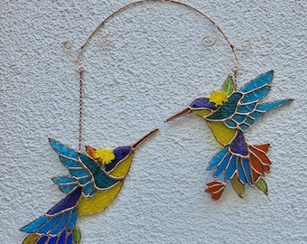 May include: Two stained glass hummingbird decorations. The hummingbirds have blue, yellow, purple, and orange wings and bodies. They are suspended from a decorative copper-coloured metal arch and chain.