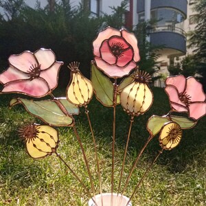 K&ouml;nnte beinhalten: Eine Ansammlung handgefertigter Glasblumen, darunter rosa Mohnblumen und gelbe Samenkapseln, mit Stielen aus Kupferdraht. Die Blumen aus durchscheinendem Glas sind in einer wei&szlig;en Vase vor gr&uuml;nem Gras angeordnet.