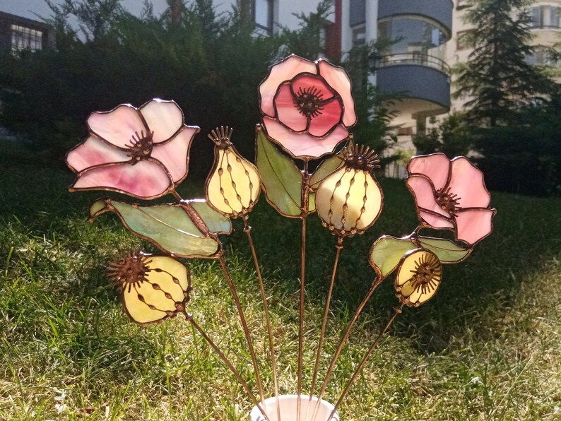 K&ouml;nnte beinhalten: Eine Ansammlung handgefertigter Glasblumen, darunter rosa Mohnblumen und gelbe Samenkapseln, mit Stielen aus Kupferdraht. Die Blumen aus durchscheinendem Glas sind in einer wei&szlig;en Vase vor gr&uuml;nem Gras angeordnet.