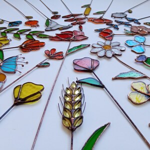 May include: A collection of colorful, stained glass garden stakes in various floral and leaf designs. The stakes feature a variety of colors, including red, yellow, blue, and green, with copper wire stems. The arrangement is on a white surface.