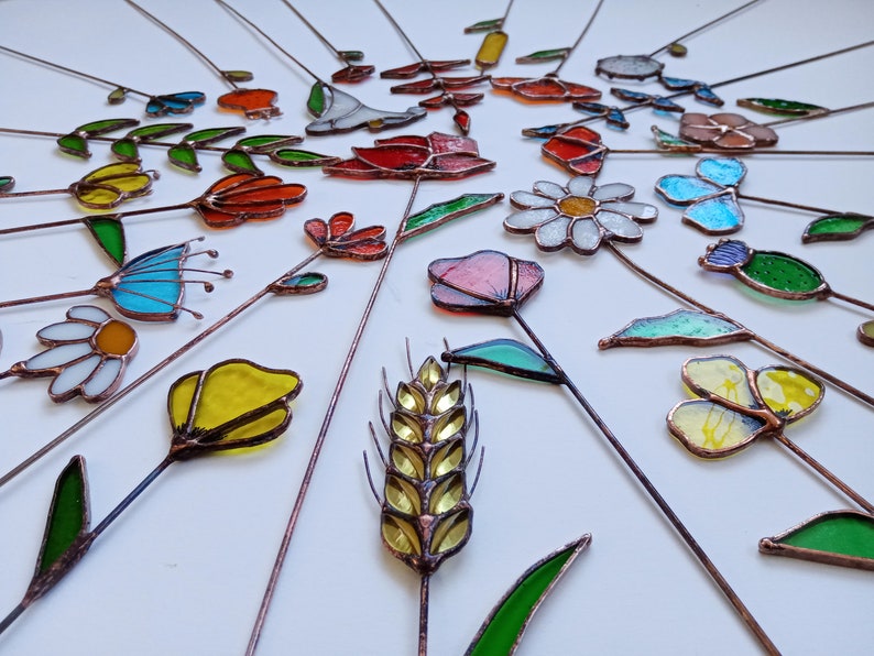 May include: A collection of colorful, stained glass garden stakes in various floral and leaf designs. The stakes feature a variety of colors, including red, yellow, blue, and green, with copper wire stems. The arrangement is on a white surface.
