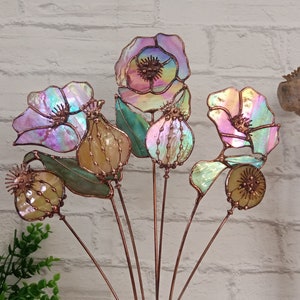 K&ouml;nnte beinhalten: Eine Sammlung von Gartensteckern aus schillerndem Glas mit Blumenmotiven. Die Stecker zeigen verschiedene Blumen- und Samenkapsel-Designs mit Kupferdrahtakzenten. Das Glas hat einen farbenfrohen, schimmernden Effekt mit Farbt&ouml;nen von Rosa, Lila und Gr&uuml;n.