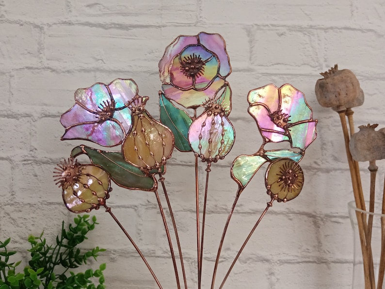 K&ouml;nnte beinhalten: Eine Sammlung von Gartensteckern aus schillerndem Glas mit Blumenmotiven. Die Stecker zeigen verschiedene Blumen- und Samenkapsel-Designs mit Kupferdrahtakzenten. Das Glas hat einen farbenfrohen, schimmernden Effekt mit Farbt&ouml;nen von Rosa, Lila und Gr&uuml;n.