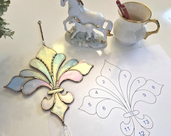 May include: A stained glass fleur-de-lis ornament in pastel iridescent colours with a clear crystal drop. A white porcelain horse figurine and a small teacup with gold trim are also present. A paper with a fleur-de-lis pattern is next to the ornament.