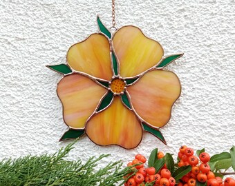 May include: A stained glass flower with yellow and peach petals, outlined in copper and accented with green leaves. The flower is hanging from a chain. The background is a white textured wall. Decorative floral art.
