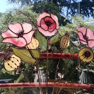 K&ouml;nnte beinhalten: Eine Sammlung von Glasmalereibl&uuml;ten in Rosa, Gelb und Gr&uuml;n. Die Blumen weisen aufwendige Details auf und sind auf Metallstielen montiert. Der Hintergrund zeigt eine verschwommene Ansicht von Gr&uuml;n und ein rotes Gel&auml;nder.