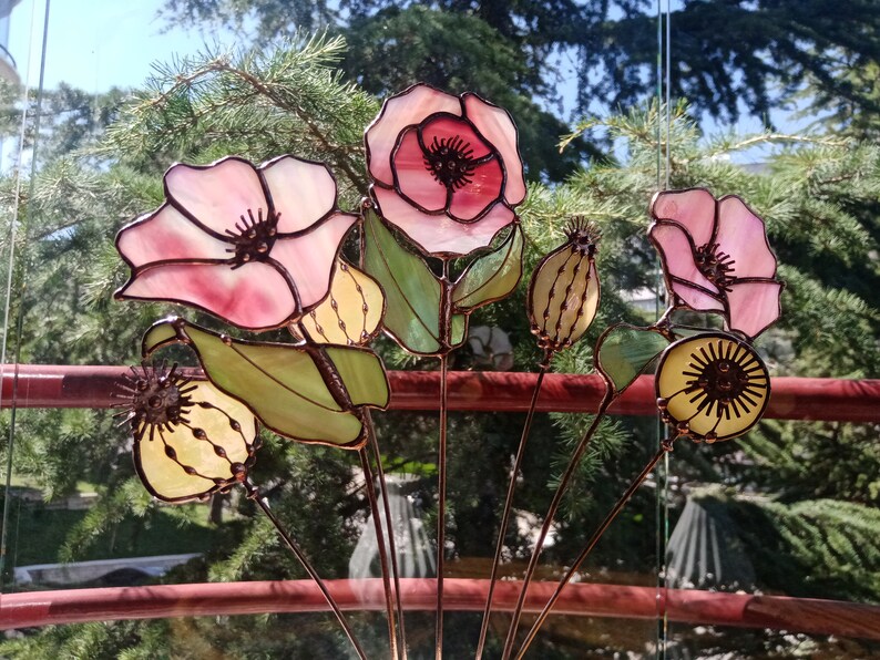 K&ouml;nnte beinhalten: Eine Sammlung von Glasmalereibl&uuml;ten in Rosa, Gelb und Gr&uuml;n. Die Blumen weisen aufwendige Details auf und sind auf Metallstielen montiert. Der Hintergrund zeigt eine verschwommene Ansicht von Gr&uuml;n und ein rotes Gel&auml;nder.