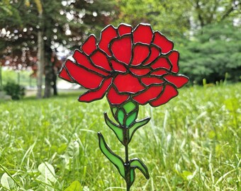 May include: A red stained glass flower garden stake with a black outline. The flower has multiple petals and green leaves. The garden stake is in a grassy area with trees in the background.