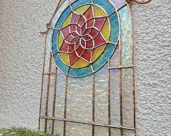 May include: Stained glass window panel with a colourful floral design. The panel features a central flower in red, yellow, and blue, surrounded by a turquoise and iridescent border. The panel is framed with copper and has a hanging leather cord.
