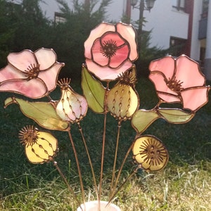 K&ouml;nnte beinhalten: Ein Glasmalerei-Blumenarrangement mit rosa und wei&szlig;en Mohnblumen, gr&uuml;nen Bl&auml;ttern und gelben Samenkapseln. Die Blumen haben ein detailliertes Design mit kupferfarbenen Metallakzenten. Das Arrangement befindet sich vor einem verschwommenen gr&uuml;nen Hintergrund.