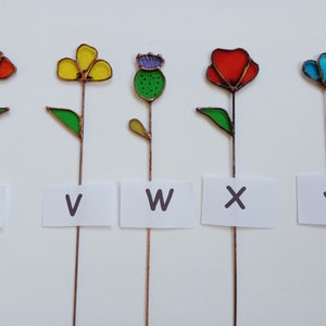 May include: Five colorful stained glass flower garden stakes. Each flower has a different color, including red, yellow, blue, and green. Each stake has a letter from the alphabet on a white label: U, V, W, X, and Y.