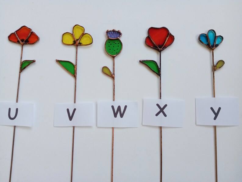 May include: Five colorful stained glass flower garden stakes. Each flower has a different color, including red, yellow, blue, and green. Each stake has a letter from the alphabet on a white label: U, V, W, X, and Y.