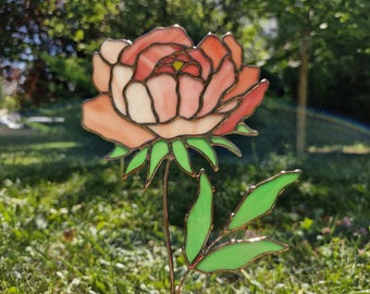 May include: A stained glass peony garden stake. The flower has peach and white petals with a yellow centre, green leaves, and a brown stem. The artwork is set against a blurred green background.