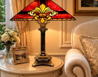 May include: A table lamp featuring a rectangular stained glass shade with a red and yellow fleur-de-lis pattern. The lamp has a dark, fluted metal column base with a square pedestal, providing a structured support for the illuminated glass top.