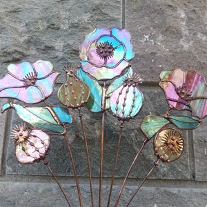 K&ouml;nnte beinhalten: Eine Sammlung von Gartensteckern aus schillerndem Glas in Form von Blumen. Die Blumen haben eine Mischung aus Farben, darunter Rosa, Blau und Gr&uuml;n, mit Kupferdrahtstielen. Das Design umfasst mohnartige Bl&uuml;ten, Samenkapseln und Bl&auml;tter.