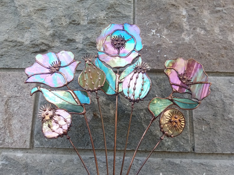 K&ouml;nnte beinhalten: Eine Sammlung von Gartensteckern aus schillerndem Glas in Form von Blumen. Die Blumen haben eine Mischung aus Farben, darunter Rosa, Blau und Gr&uuml;n, mit Kupferdrahtstielen. Das Design umfasst mohnartige Bl&uuml;ten, Samenkapseln und Bl&auml;tter.
