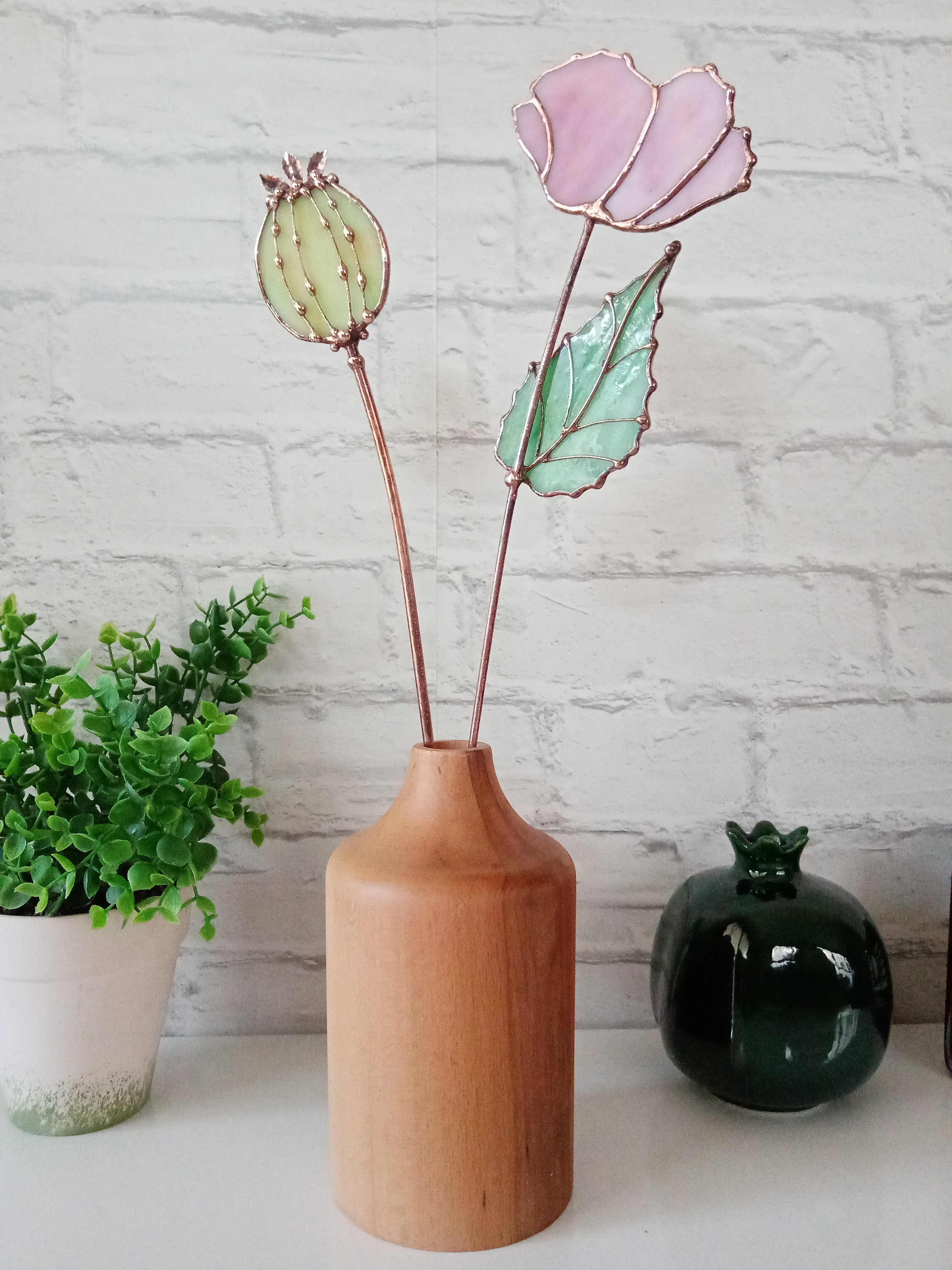 May include: A wooden vase with a light brown finish holds three stained glass flowers. The flowers are a pink poppy, a green leaf, and a green poppy bud.