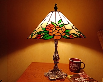 May include: A decorative table lamp in the Tiffany style, featuring a stained glass shade with a floral pattern of orange and red roses and green leaves. The lamp has a bronze base. A red mug and a book are on the table.