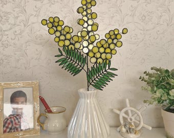 May include: A stained glass mimosa flower sculpture featuring bright yellow circular glass pieces arranged in clusters on thin metal stems with green glass leaves, standing upright in a white ribbed ceramic vase with a tapered neck.