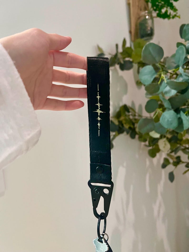 K&ouml;nnte beinhalten: Ein schwarzes Schl&uuml;sselband aus Stoff mit einer mittigen, hellen Schallwellengrafik. Das Band ist mit einem schwarzen Metalldruckknopf gesichert und verf&uuml;gt &uuml;ber einen schwarzen Metallkarabiner am unteren Ende zum Befestigen von Schl&uuml;sseln.
