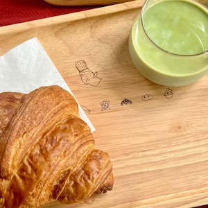 Könnte beinhalten: Ein Holztablett mit einem Croissant auf einer Serviette und einem Glas grüner Matcha Latte. Das Tablett hat eine helle Holzmaserung und abgerundete Kanten. Der Hintergrund ist eine rote Oberfläche.