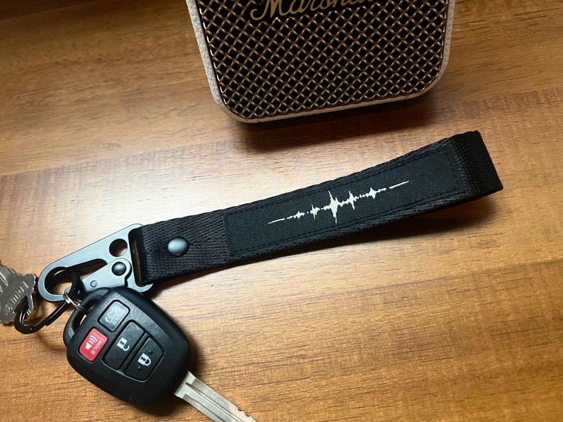 K&ouml;nnte beinhalten: Ein schwarzes Schl&uuml;sselband aus Stoff mit einem wei&szlig;en Schallwellen-Grafikdruck. Das Band ist an einem schwarzen Metallkarabiner befestigt, an dem sich ein schwarzer Autoschl&uuml;ssel aus Kunststoff mit vier Tasten und einem Metallschl&uuml;sselbart befindet.