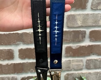 May include: Two wrist lanyards made of woven fabric, one black and one navy blue. Each features a rectangular patch with a white sound wave graphic printed vertically. The black lanyard has a matte black metal clasp, while the navy blue lanyard has a silver metal clasp.