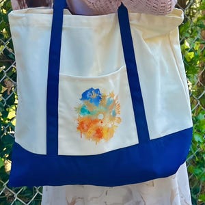 Könnte beinhalten: Cremefarbene Tragetasche mit marineblauem Boden und Trägern. Die Vordertasche zeigt ein farbenfrohes abstraktes Blumendesign in Blau-, Orange- und Gelbtönen. Die Tasche besteht aus einem robusten Material und eignet sich zum Tragen verschiedener Gegenstände.