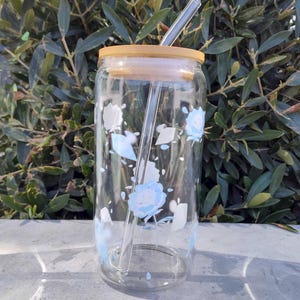 Könnte beinhalten: Eine wiederverwendbare Wasserflasche aus klarem Glas mit einem Holzdeckel und einem Glasstrohhalm. Die Flasche hat ein Blumenmuster mit blauen und weißen Blüten.