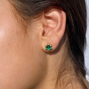 Può includere: Un paio di orecchini a bottone dorati con una pietra preziosa verde al centro. Gli orecchini hanno la forma di un fiore con piccole perle dorate intorno al bordo.