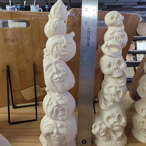 Peut inclure: Deux sculptures en céramique blanche à côté d'une règle en métal Westcott. La sculpture de gauche est une pile de cinq citrouilles sculptées avec des visages, et celle de droite est une pile de cinq crânes. La règle indique 33 cm.