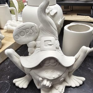 May include: An unpainted white ceramic figurine of a frog wearing a tall, pointed hat with a belt and buckle. The frog has a hand to its chin and is looking at an open book. A speech bubble attached to the hat features the embossed text OOPS!