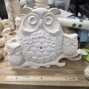 May include: A white ceramic owl clock figurine featuring a large central owl with numbers 1 through 12 and two smaller owls on either side, all perched on a branch. A wooden ruler placed in front shows the piece is approximately 12 inches wide.