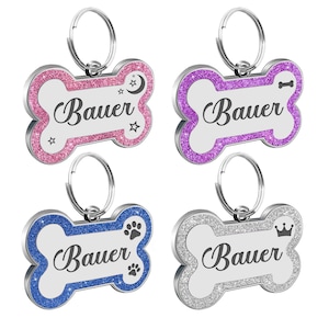 May include: Four silver bone-shaped dog tags with different colored glitter borders. The tags have the name "Bauer" engraved on them and each has a different design: a crescent moon and stars, a bone, a paw print, and a crown.