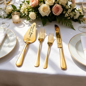 May include: A gold-colored wedding cake server, two forks, and a knife set on a white tablecloth. The server and knife are engraved with names and a date. A floral arrangement of white and pink roses is in the background.