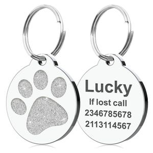 Op de afbeelding: Twee ronde, zilveren huisdier-ID-tags. E&eacute;n tag heeft een glitterende pootafdruk. De andere tag heeft het woord "Lucky" en twee telefoonnummers. Beide tags hebben een zilveren ring voor bevestiging.