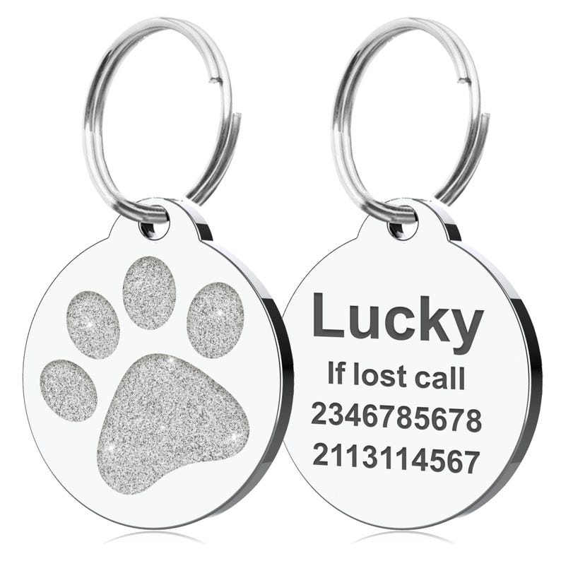 Op de afbeelding: Twee ronde, zilveren huisdier-ID-tags. E&eacute;n tag heeft een glitterende pootafdruk. De andere tag heeft het woord "Lucky" en twee telefoonnummers. Beide tags hebben een zilveren ring voor bevestiging.