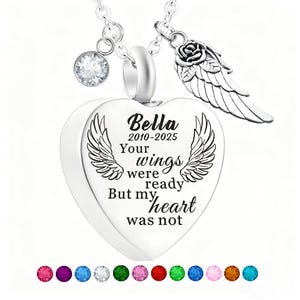 May include: A silver heart-shaped pendant necklace with the name "Bella" and dates 2010-2025 engraved on it. The pendant has a pair of wings and the words "Your wings were ready But my heart was not". A crystal and a rose with wings charm are attached.