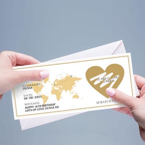 May include: A gold and white scratch-off card with a world map design and the text "We are going to Paris." The card includes the passenger's name, date, and a birthday message. The card is held in front of a light gray background.