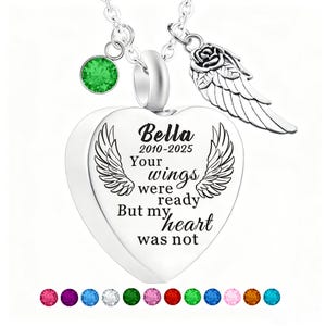 May include: A silver heart-shaped pendant necklace with the inscription "Bella 2010-2025 Your wings were ready But my heart was not." The necklace includes a green gemstone, a rose and wing charm, and a selection of colorful gemstones.
